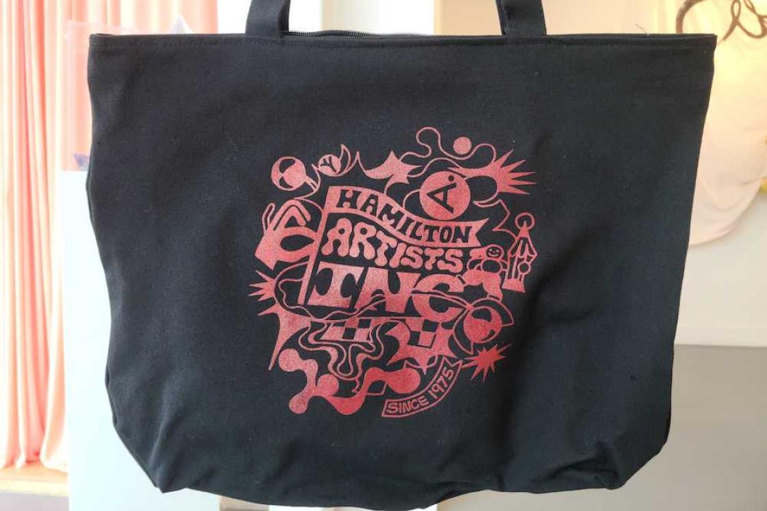 The image is a photo of a black tote bag with a monochromatic red design. The design is a surreal rendering of the Hamilton Artists Inc logo, with abstract shapes and patterns exploding from the centre.