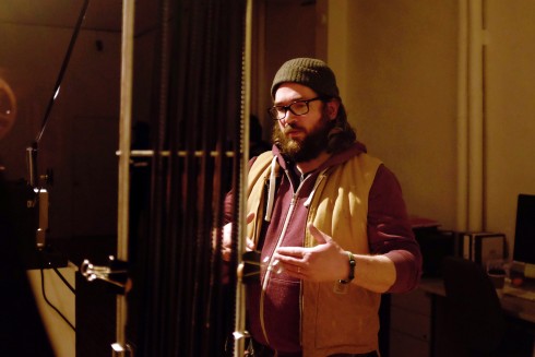 Derek is standing in a dimly-lit room, gesturing towards a contraption hanging directly in front of him. Derek has long brown hair and beard, and is wearing black-rimmed glasses, a green toque, burgundy hoodie, and sandy-coloured vest.