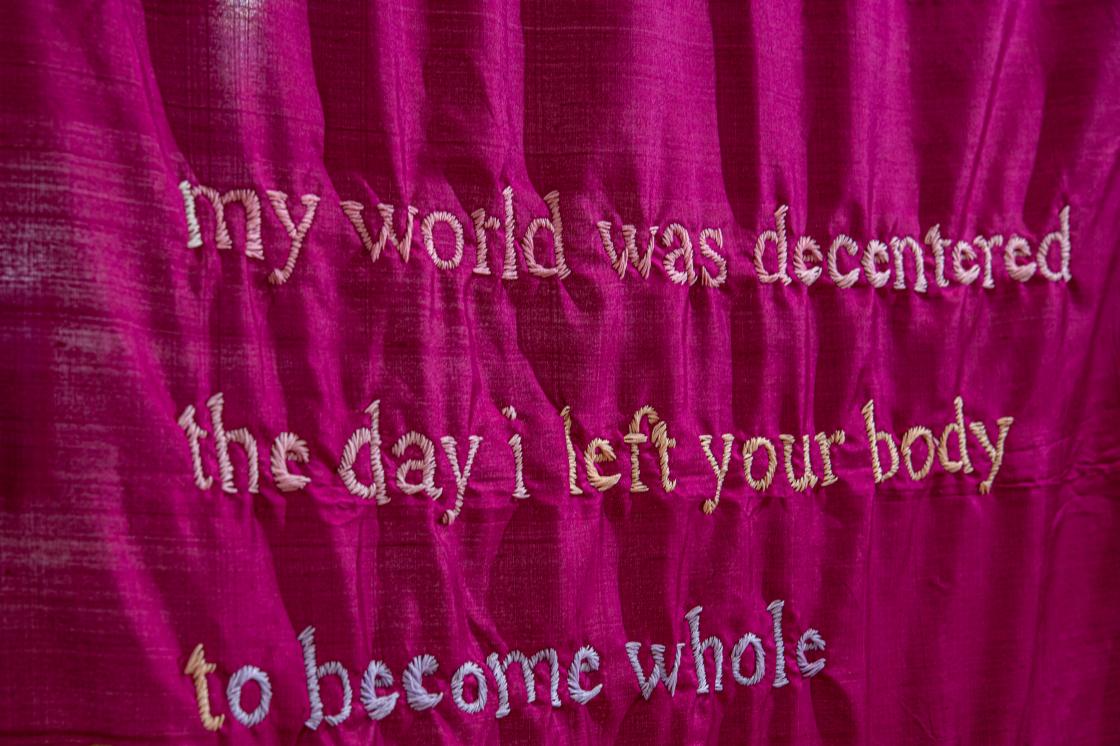 A detailed image of a hot pink saree by Par Nair with hand embroidered text which says "my world was decentered the day i left your body to become whole"