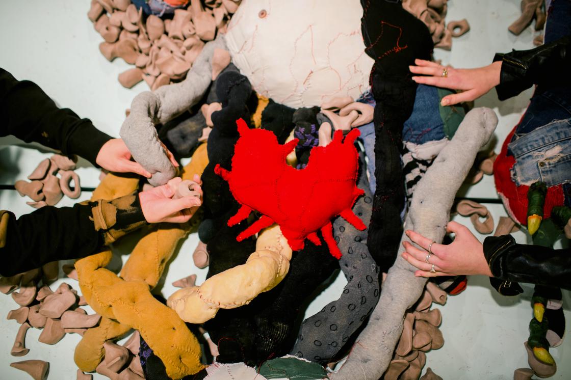 Two light-skinned people reach out to touch Big Softie’s guts, which are soft and dimpled and made from stuffed knee socks and nylon stockings. Many small, organically shaped unfired clay objects are scattered amongst the textile-based viscera. At the centre of it all lies Big Softie’s heart, a bright red patchwork soft sculpture with tendrils extending outward.