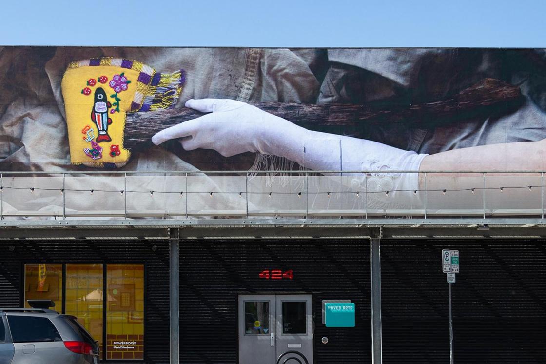 The image is a photo of PAVED Arts. The front of the gallery features a billboard. The billboard features a photograph of a gloved arm holding the handle of an axe. The head of the axe features a sheath that has been decorated with Western North American Indigenous-inspired leatherwork and beadwork.