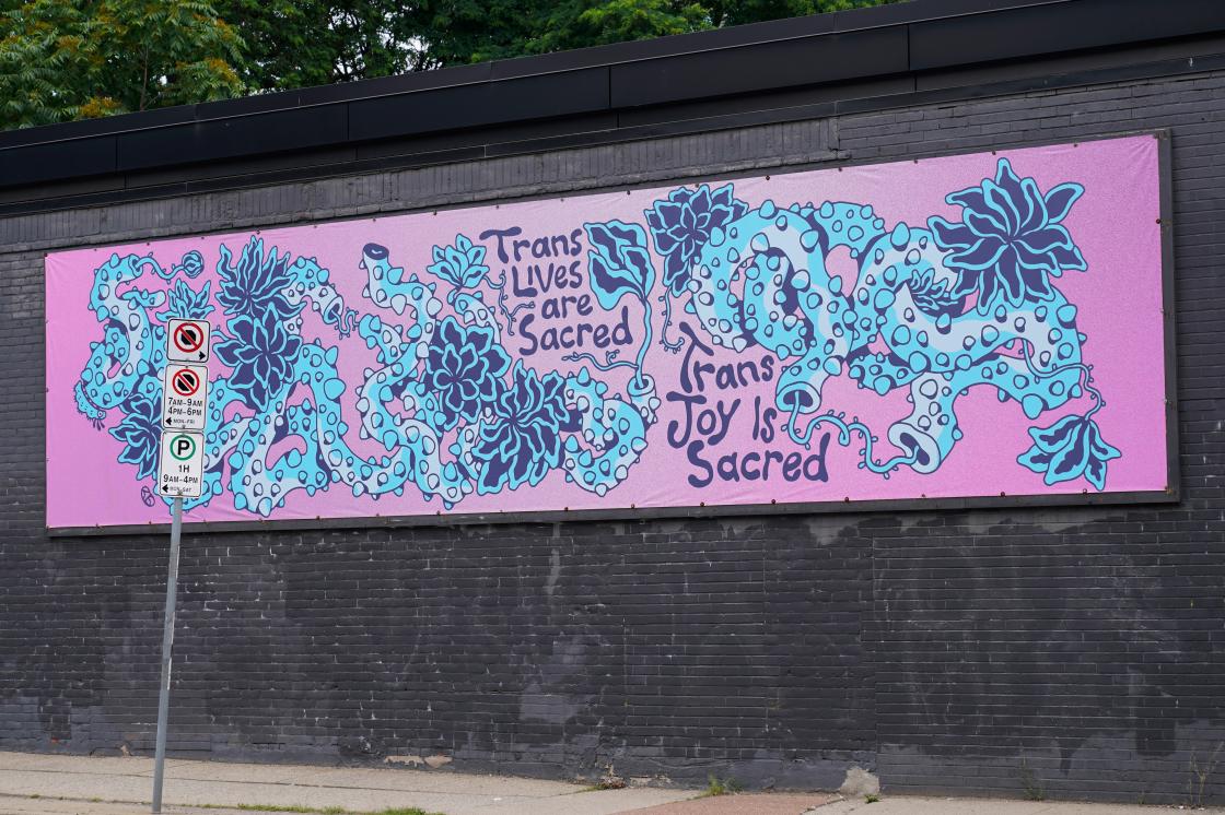 A rectangular purple and blue illustrated banner installed on a gray brick wall on the exterior of Hamilton Artists Inc. The banner is a drawn depiction of blue-coloured vines with thorns intertwined with each other  across a wide background of a purple gradient. The vines have light blue highlights and flowers and smaller slender stems are interspersed amongst the coils of the vines. The text “Trans Lives are Sacred'' and “Trans Joy is Sacred” is nestled between the vines in the middle of the illustration.