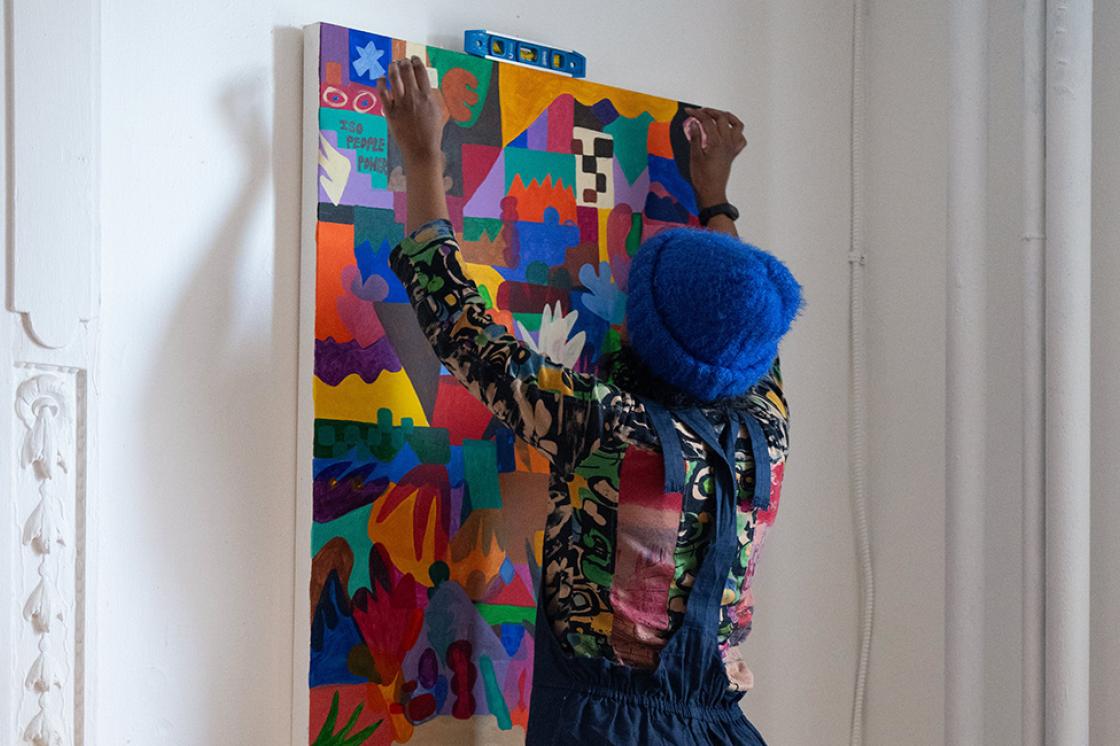 Sahra, a black non-binary person with a blue hat a patterned shirt and denim overalls levels a colourful abstract painting on a white wall