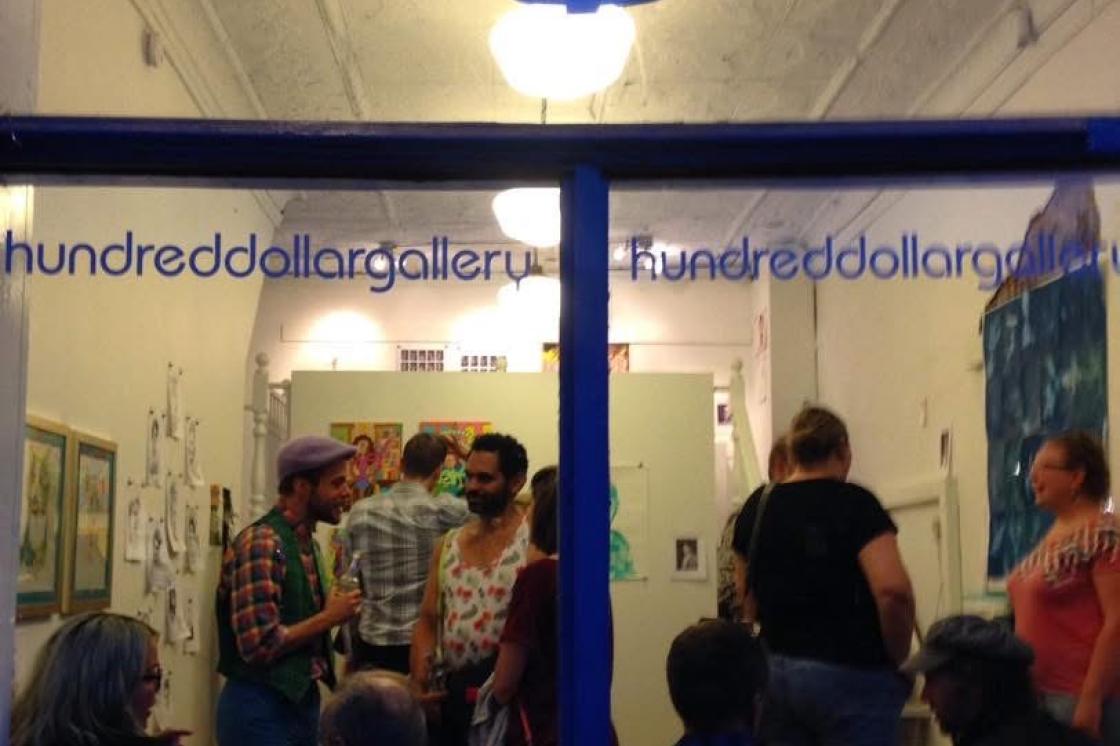 This is an image of the Hundred Dollar Gallery that was located on James Street North, and the photo was taken some time in the 2010s. The photo was taken from outside of the window, looking inwards at people who are chatting amongst themselves in the space. There is a large blue "$" sign on the top part of the window, and the window also says "hundreddollargallery" twice in blue text.