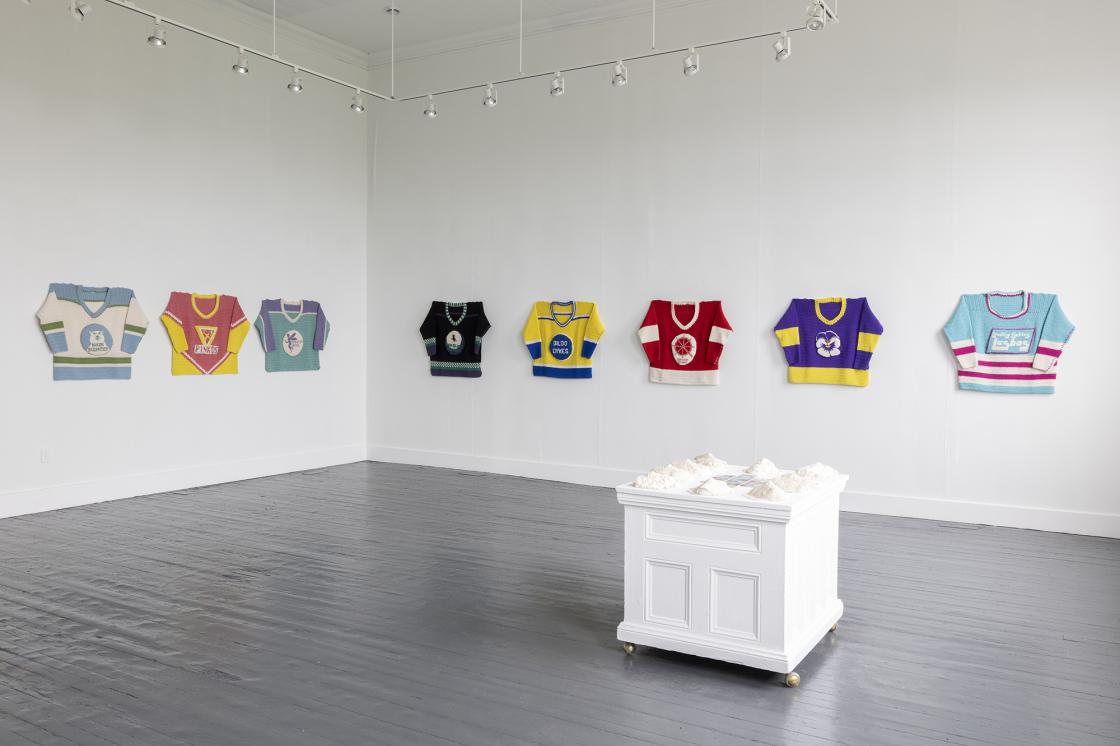 This is an image from Lucas Morneau's exhibition "Queer Newfoundland Hockey League", taken in a gallery space. It features 8 crocheted hockey jerseys along two intersecting walls, and a plinth with crocheted masks in the middle of the space.