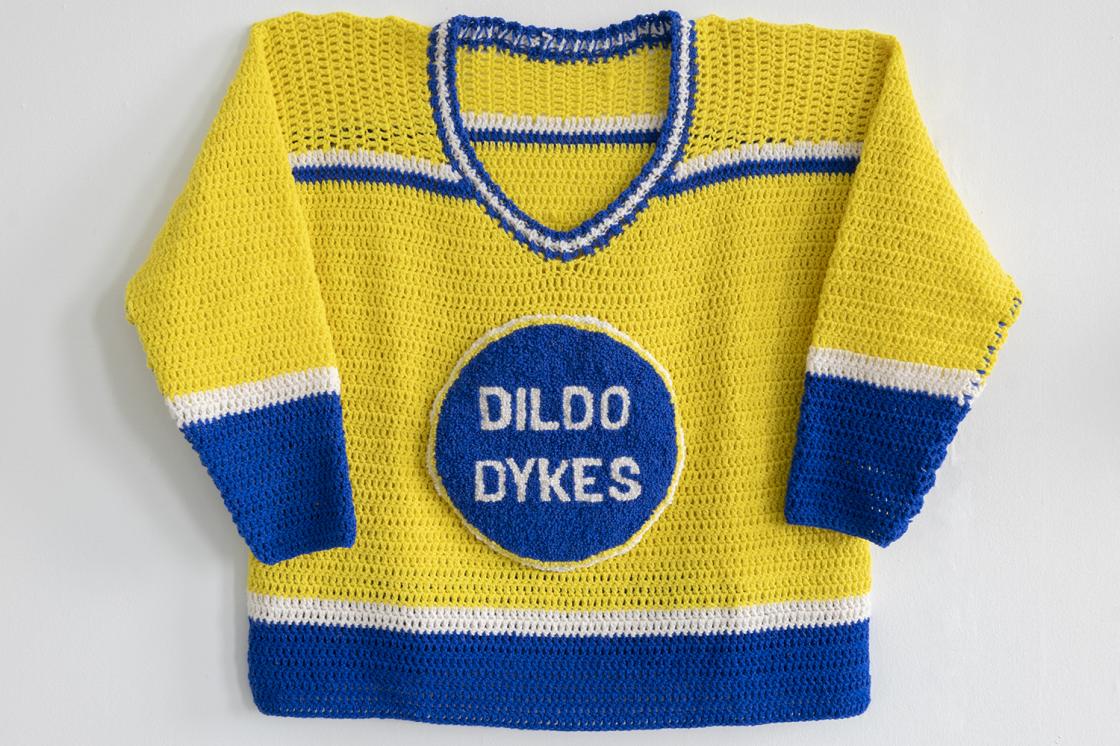 A crocheted hockey jersey for Morneau's exhibit, "Queer Newfoundland Hockey League". It is crocheted with yellow as the dominant team colour, with white and blue stripe accents on the sleeves and the body. The team crest says "Dildo Dykes" in a blue circle in the middle of the jersey.