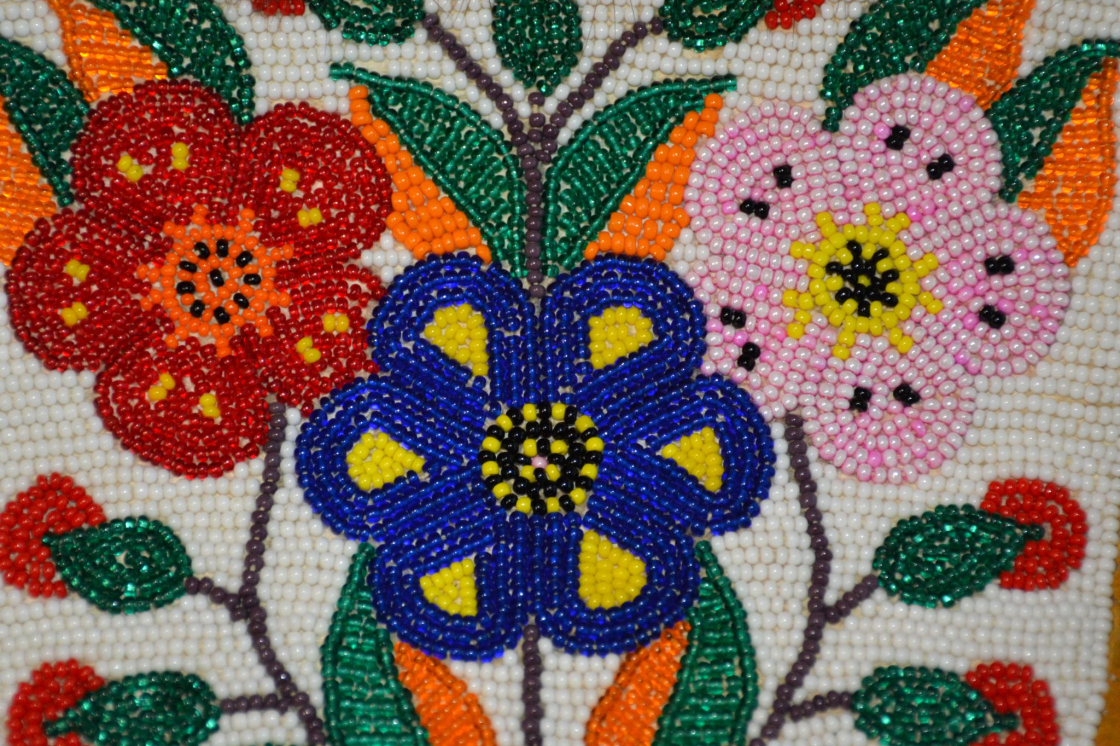 A close-up shot of beaded work, featuring a red flower on the left, a royal blue flower in the lower centre, and a light pink flower on the right. There are deep emerald green beaded leaves with orange details, along with a white background on the piece. 