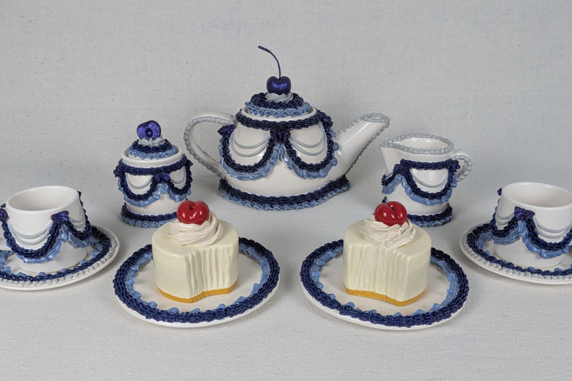 A collection of ceramic works by Solange Roy. They show teacups, teapots, a sugar and creamer all decorated with blue and white icing. The teapot has a blue cherry on top. There are also two pieces of ceramic cake in shades of vanilla.