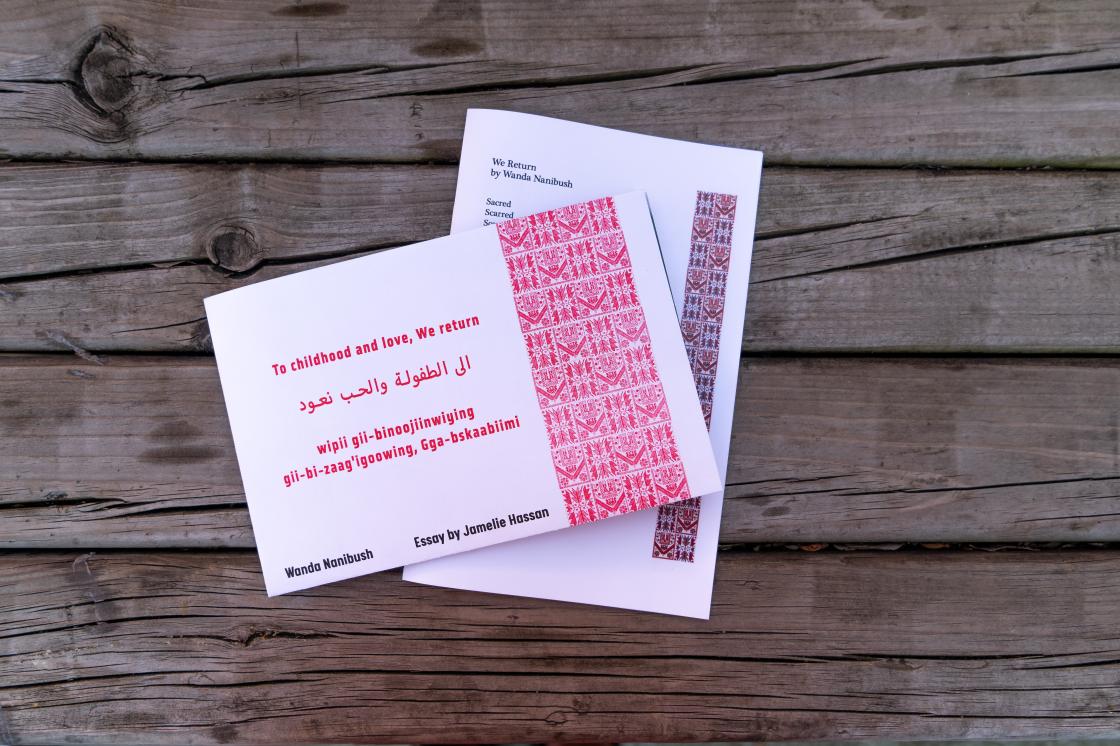 A bird’s eye view of two white paper rectangular folded publications with red detailing sitting interconnected on a wood bench. The publication on the top reads: to childhood with love, We return in red text, followed by the Arabic and Anishinaabemowin translation of the same title. In black text on the bottom reads: Wanda Nanibush, Essay by Jamelie Hassan. To the right is a red Palestinian textile pattern. The publication underneath features poetry by Wanda, with a similar red Palestinian textile pattern.