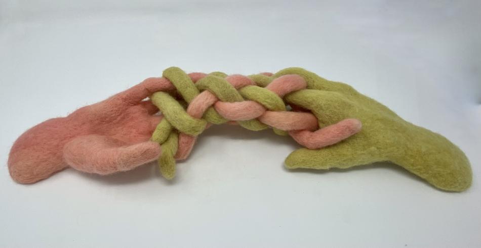 A wool felted sculpture by Alana Morouney which features two life sized hands with exaugurated fingers intertwined together. One hand is pink and one is green.