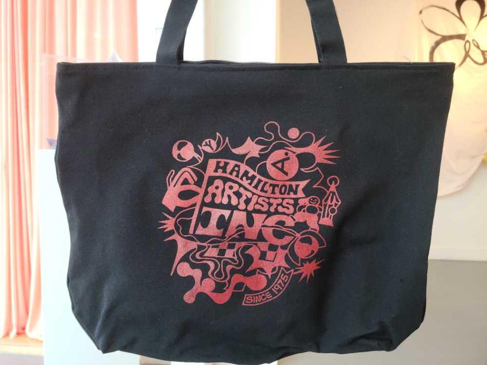 The image is a photo of a black tote bag with a monochromatic red design. The design is a surreal rendering of the Hamilton Artists Inc logo, with abstract shapes and patterns exploding from the centre.