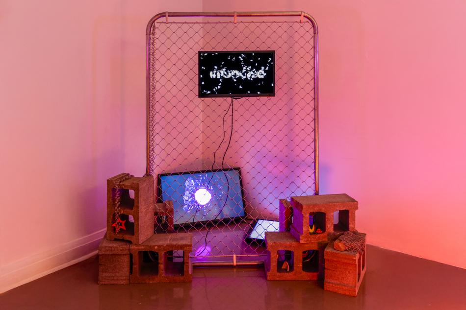 a sculptural installation in the corner of two walls, lit by pink and blue light featuring grey cinder blocks, two monitors and a panel of chainlink fencing. 