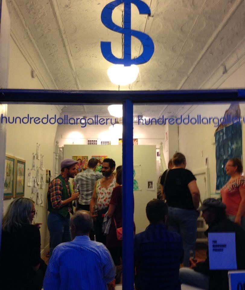 This is an image of the Hundred Dollar Gallery that was located on James Street North, and the photo was taken some time in the 2010s. The photo was taken from outside of the window, looking inwards at people who are chatting amongst themselves in the space. There is a large blue "$" sign on the top part of the window, and the window also says "hundreddollargallery" twice in blue text.