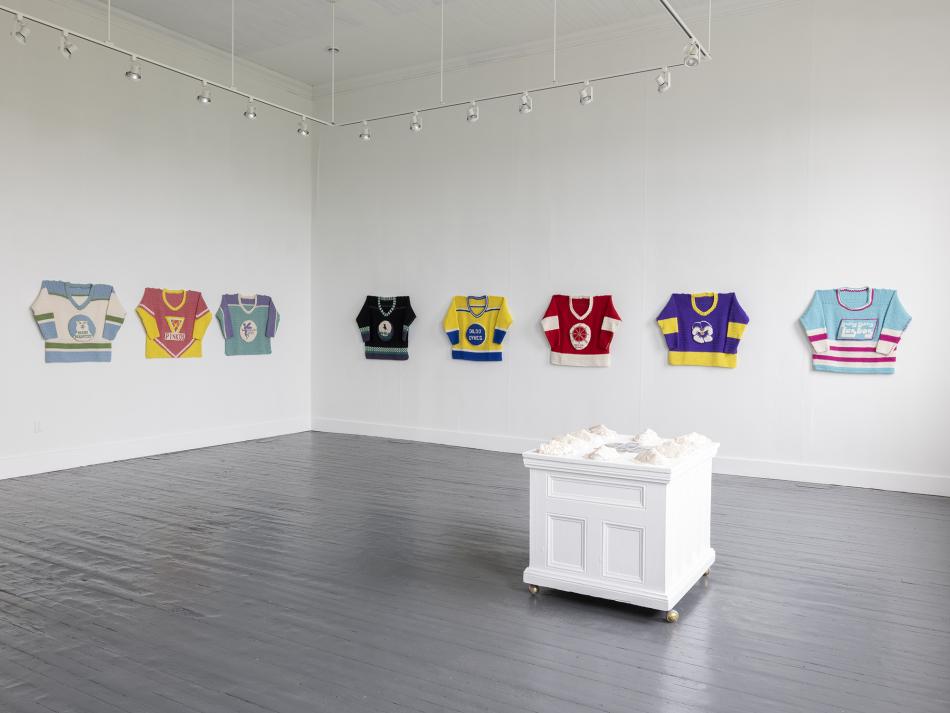 This is an image from Lucas Morneau's exhibition "Queer Newfoundland Hockey League", taken in a gallery space. It features 8 crocheted hockey jerseys along two intersecting walls, and a plinth with crocheted masks in the middle of the space.