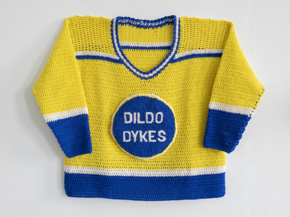 A crocheted hockey jersey for Morneau's exhibit, "Queer Newfoundland Hockey League". It is crocheted with yellow as the dominant team colour, with white and blue stripe accents on the sleeves and the body. The team crest says "Dildo Dykes" in a blue circle in the middle of the jersey. 