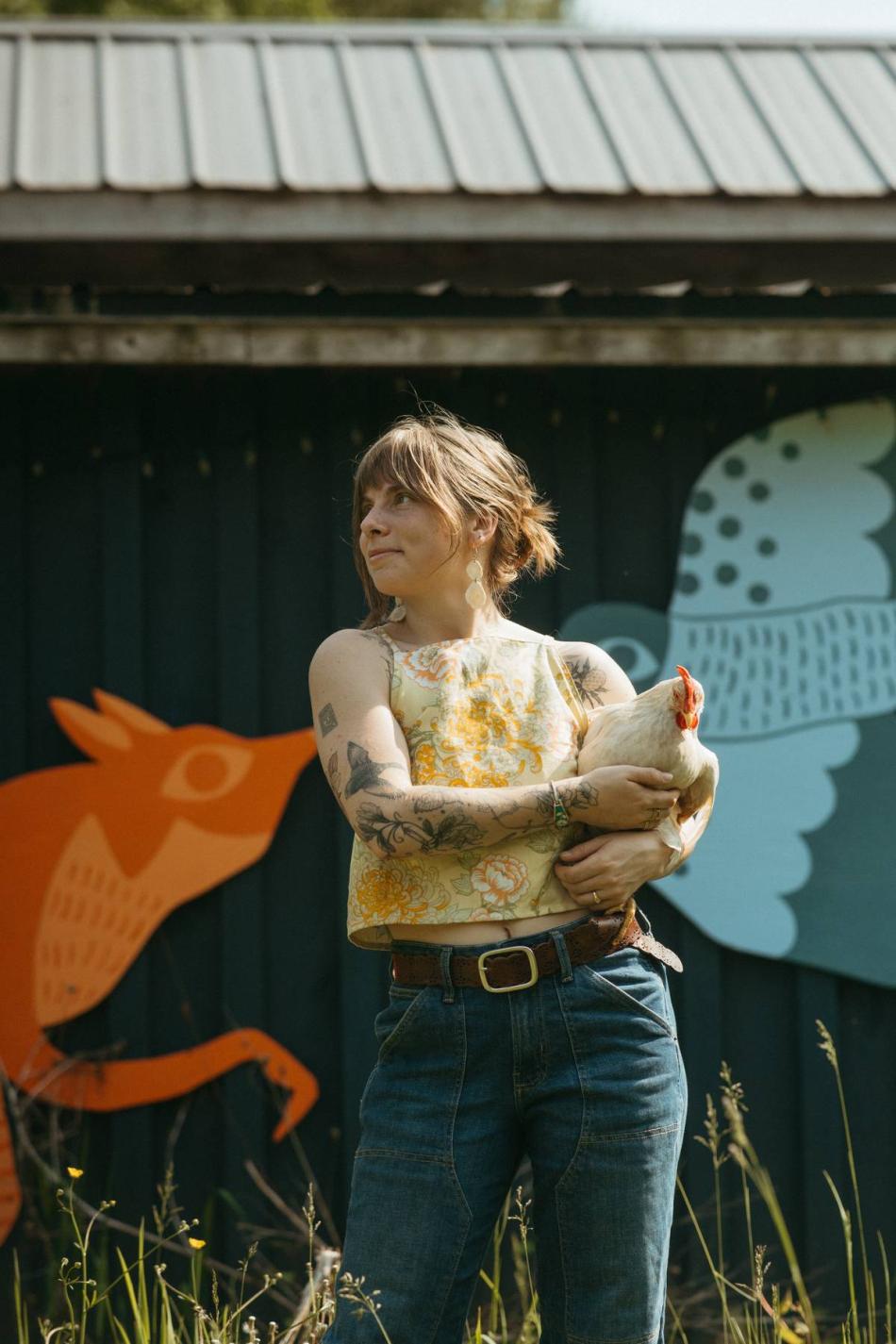 Abby is standing in front of a building with a painted mural of a fox and a bird, with a hen in her arms.