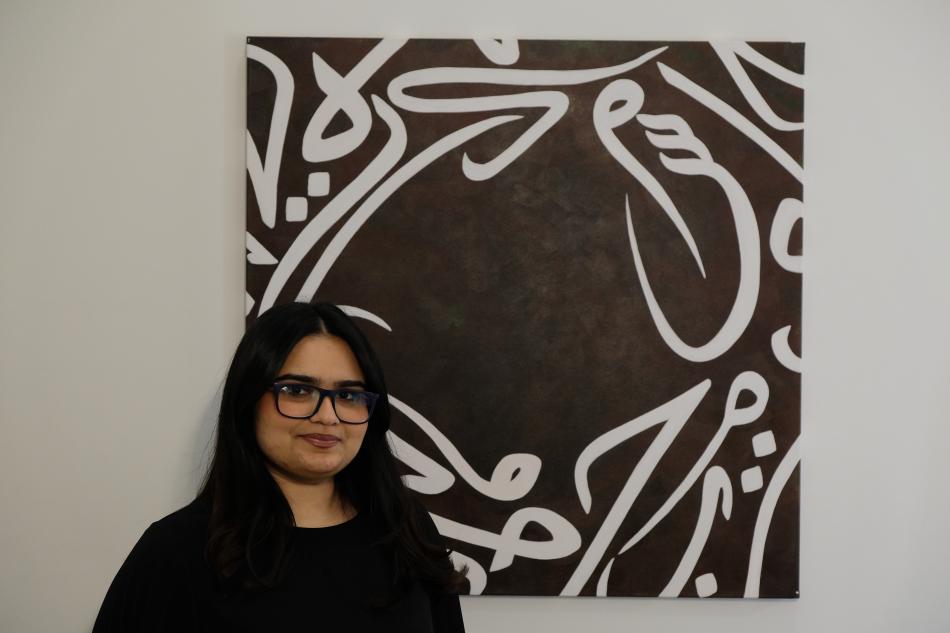 Rania is a woman with long dark hair, glasses and a black top, standing in front of one of her paintings.