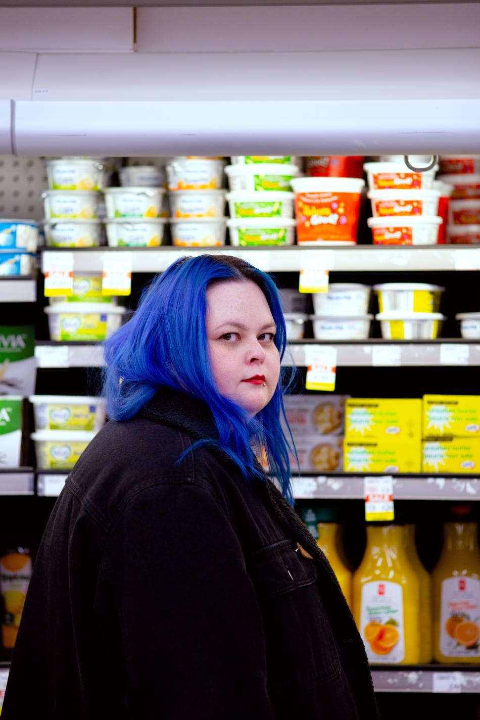 A person with long, cobalt blue hair and a black coat stands and looks at the camera sideways in front of the fridge section of a grocery store.