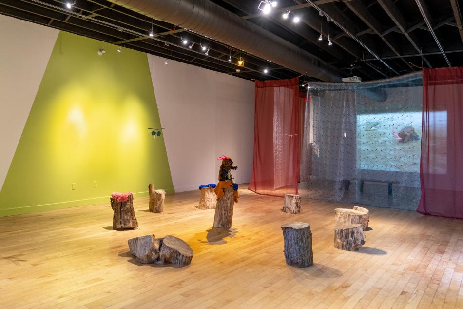 This is a photo from our exhibition, Prince Sipihko, Sailor Wapakwiniy. There's a beaver-like creature sitting on a stump surrounded by other stumps in the centre of the room. Red and light blue curtains mask a projection of a video on the far wall.