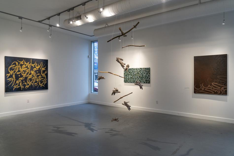 This is a photo of 'Dreams to get you through the night'. There is a mobile featuring pulp-made bunnies floating in the middle of the gallery. One painting by Rania Haider is on the left wall, and two paintings by Rania Haider are on the adjacent wall. There's a window with a blue light casting in.