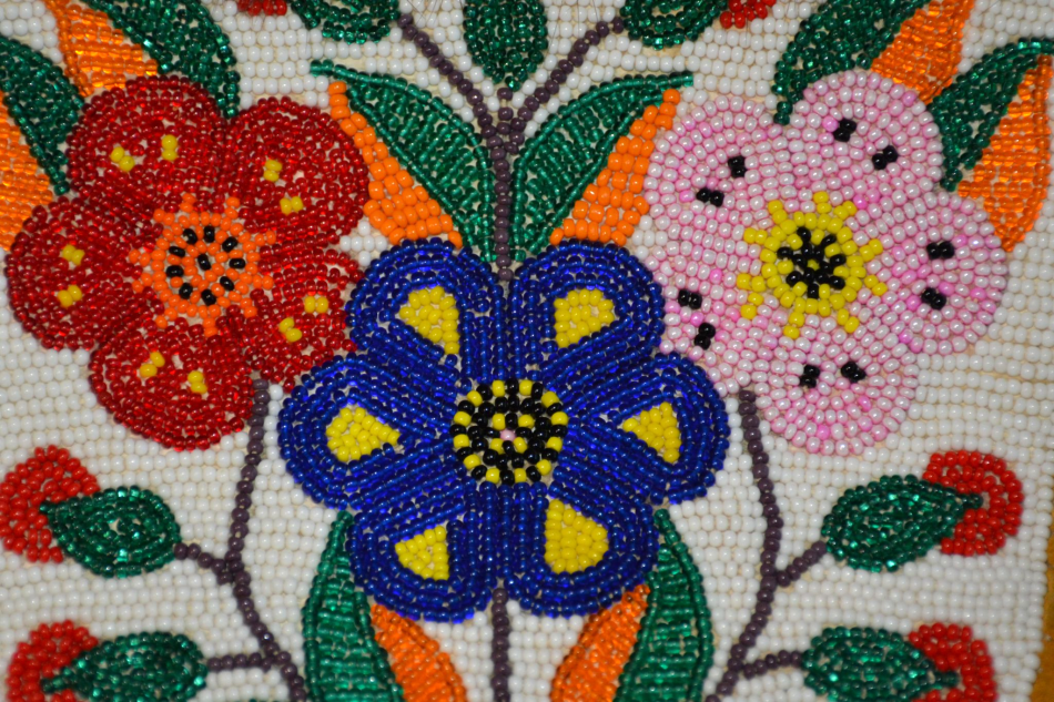 A close-up shot of beaded work, featuring a red flower on the left, a royal blue flower in the lower centre, and a light pink flower on the right. There are deep emerald green beaded leaves with orange details, along with a white background on the piece. 