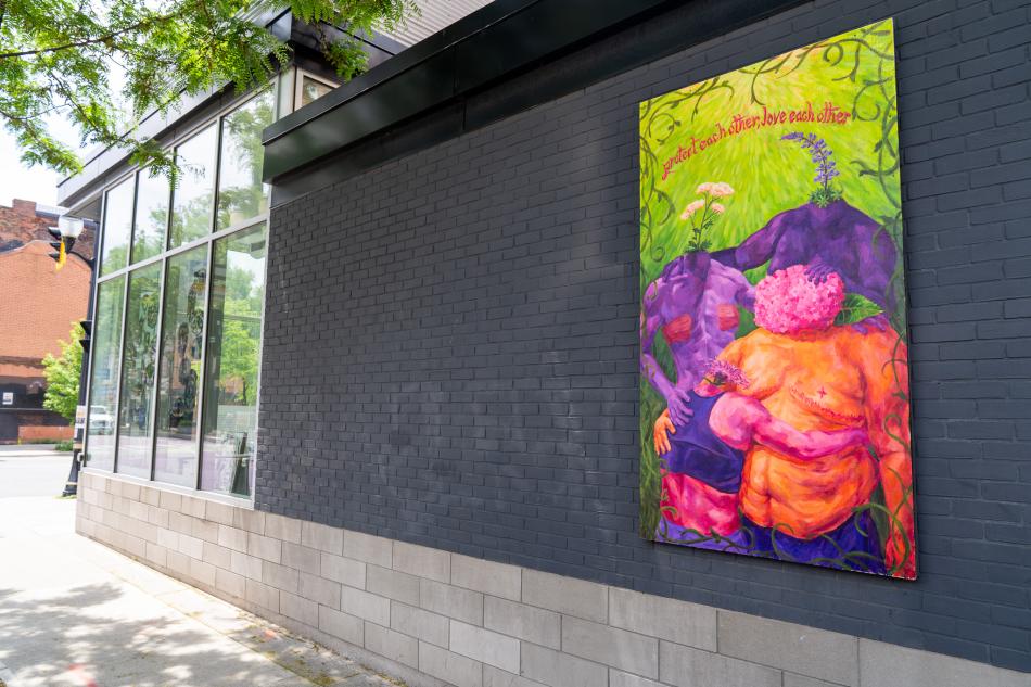 Ash Milne's billboard, "Bloom", is on a grey brick wall on James Street North, with glass windows to the left of the wall. The billboard is on the right side of the brick wall and in the right side of the photo. It features three colourful, headless figures hugging each other. Flowers bloom from their heads. The background is green grass, with vines painted around the top edges of the painting.