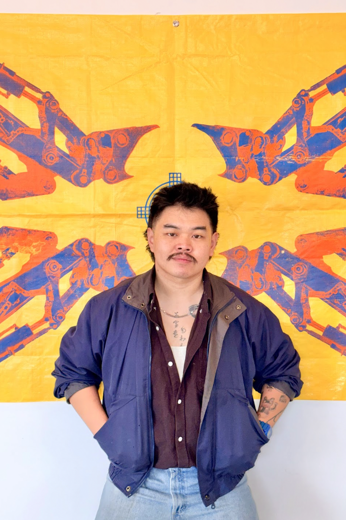 A photo of Kev Liang, who is a Chinese-Canadian man with short, dark hair, dark eyes and a moustache. He is wearing a navy blue coat over a brown button-down top, and has tattoos on his chest. Behind him is one of his tarp artworks. 