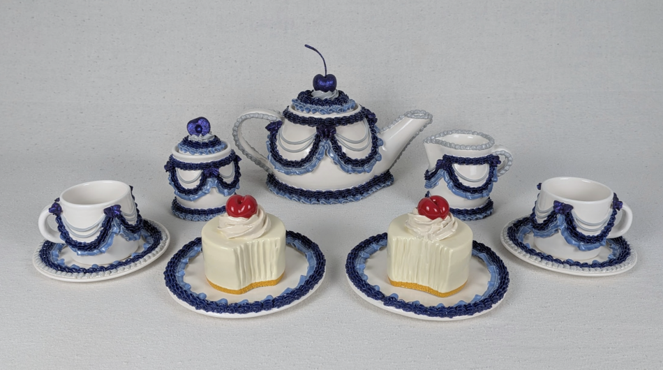 A collection of ceramic works by Solange Roy. They show teacups, teapots, a sugar and creamer all decorated with blue and white icing. The teapot has a blue cherry on top. There are also two pieces of ceramic cake in shades of vanilla.