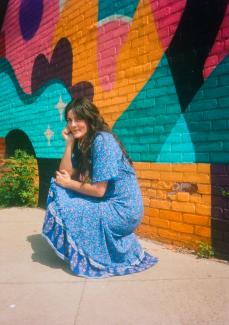 A woman in a floral blue dress with long brown hair crouches and looks toward the camera, resting her hand on her chin. There's a colourful painted brick mural behind her.