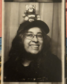 A black and white image - an analog film Paula took while they visited the Slice of Life gallery in Vancouver. They have long dark hair, are wearing a black cap and glasses. A plushie raccoon named Roberto sits on top of their hat. Paula is smiling and looking slightly left of the camera.