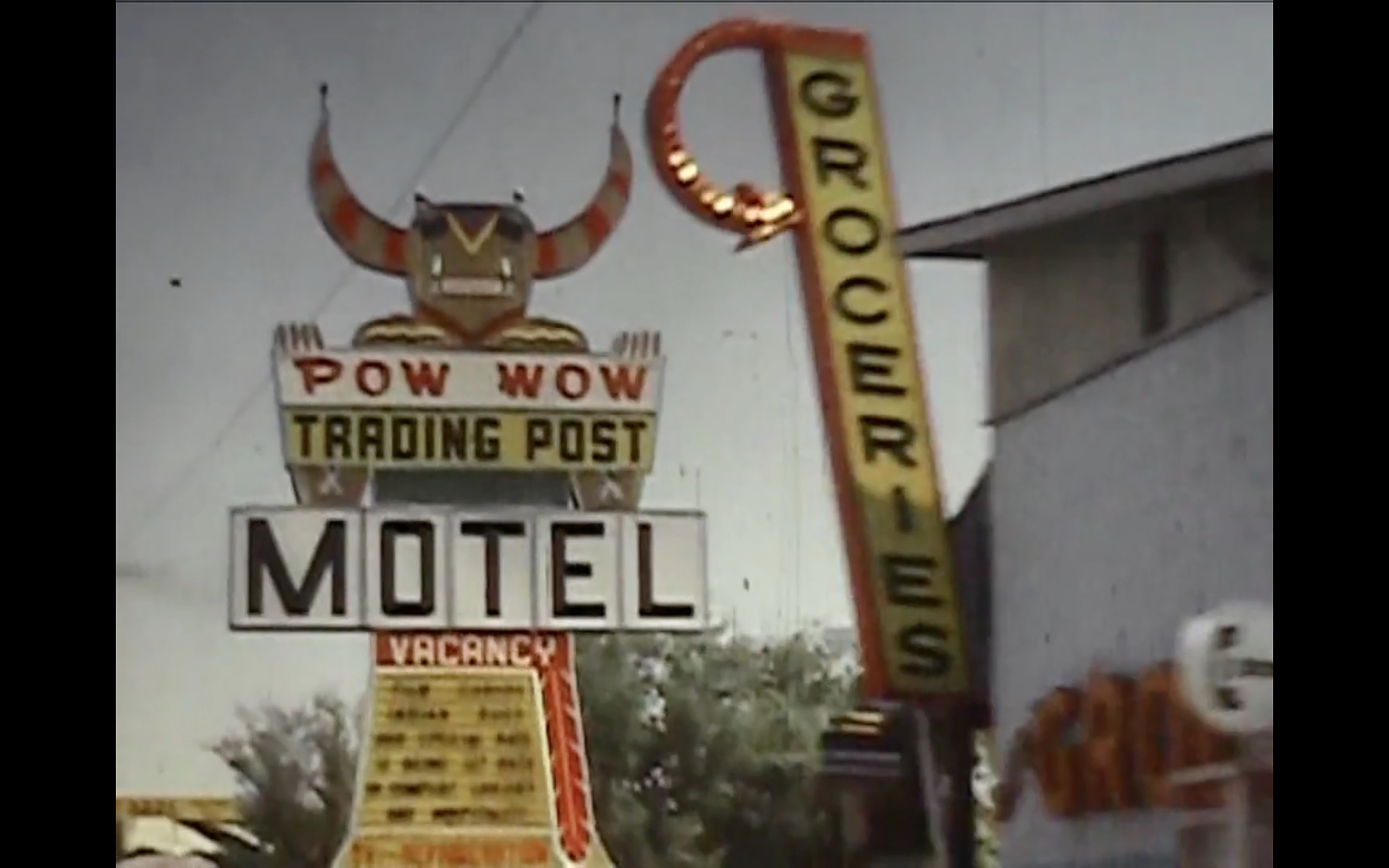 A still from the film "Class Order Family Tribe" by Rob Fatal. The still features a wide shot of outdoor signage outside of a grocery stor. The still is from a film, and the signage in focus is of a particularly old aesthetic. The leftside of the image features a sign that advertises a "Pow wow trading post" motel. A cartoon figure of a native person with horns holds up the sign for the trading post.
