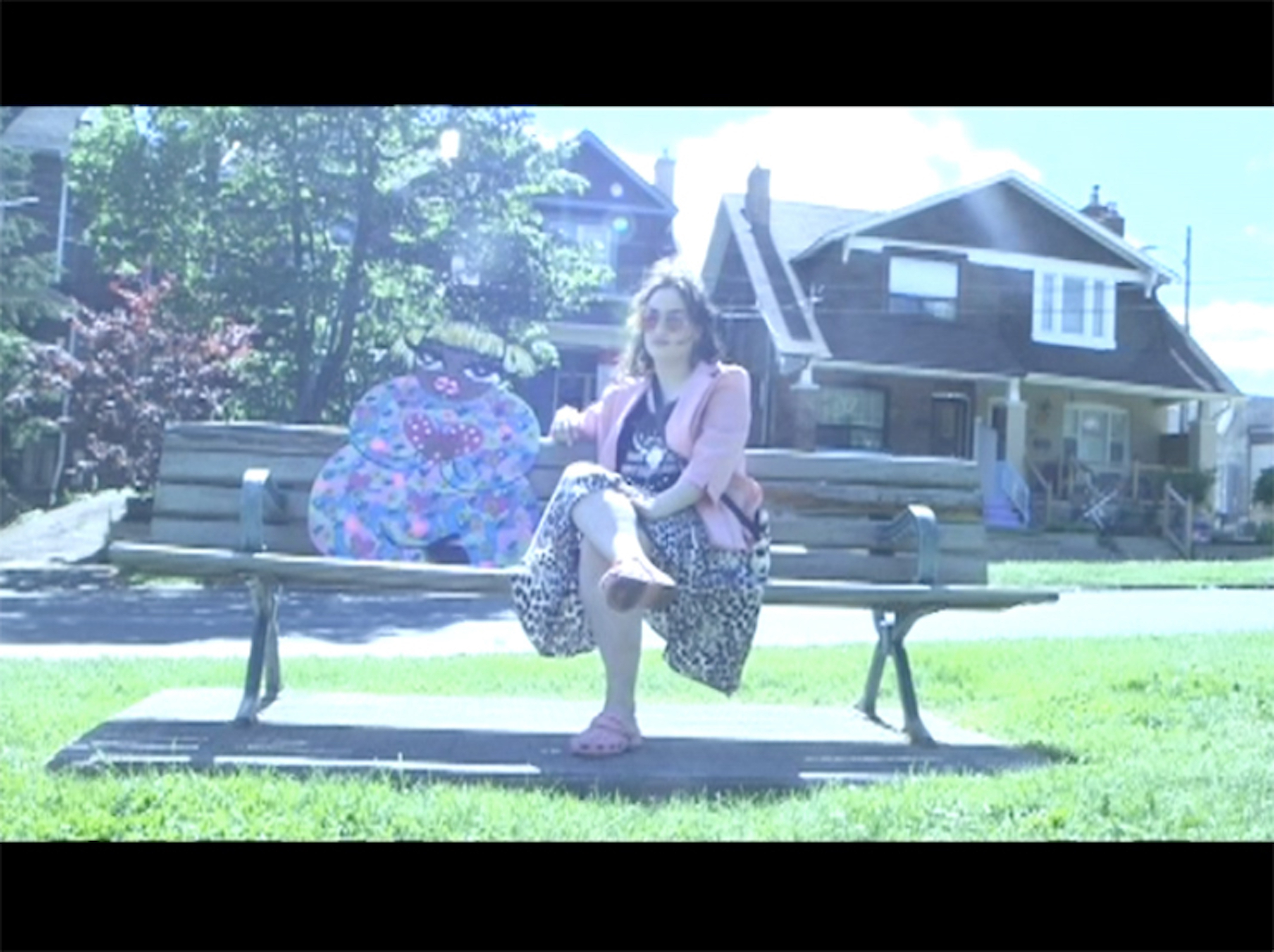 A still from the film "Activate NDN Consciousness" by Natalie King. The still features a woman sitting on a public bench alongside a two-dimensional wood caricature of a woman of color.